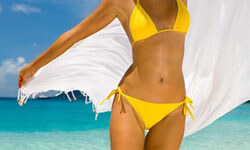 Picture of a woman, happy with her abdomen and waist liposuction procedure she had with Top Plastic Surgeons in beautiful Cancun, Mexico.  The woman is wearing a two-Dpiece yellow bathing suit and walking through the surf at the beach.