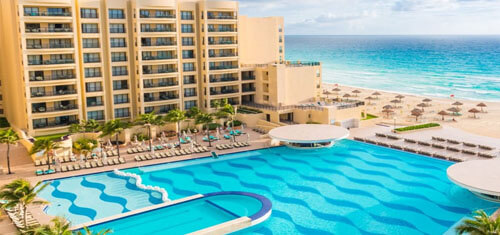 Picture of a beautiful Cancun beach with blue water and a hotel and swimming pool featured in the foreground.