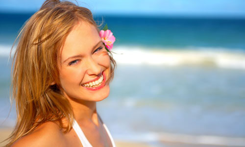 Close-up picture of a beautiful woman, happy with her face lift with neck lift in Cancun, Mexico.  The woman has long sandy blonde hair and is looking directly at the camera with the ocean behind her.
