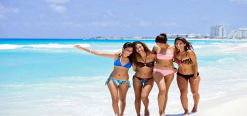 ). Picture of four women in Cancun representing the vacation opportunities available for patients having plastic surgery in Cancun, Mexico.  The picture shows four women in two piece bikini bathing suits walking on the beach.