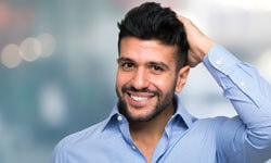 Picture of a man, happy with his hair transplant procedure he had with Top Plastic Surgeons in beautiful Cancun, Mexico.  The man is wearing a blue shirt with an open collar and holding his left hand to his head.