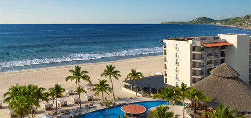 Picture of a beautiful Costa Rica beach with blue water and a hotel and swimming pool featured in the foreground.  The pool is oval shaped with palm trees all around it