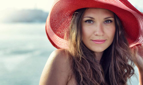 Portrait picture of a beautiful woman, happy with her face lift with neck lift procedure i in Cancun, Mexico.  The woman has long dark hair, is looking directly into the camera and is wearing a soft red beach hat highlighting her face lift with neck lift.
