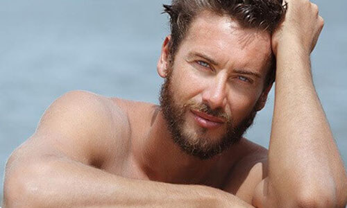 Close-up picture of a man,  happy with his hair transplant  procedure he had in Cancun, Mexico.  The man is shown leaning against the edge of a pool and looking directly at the camera with an arm raised to his head to feature his hair transplant.