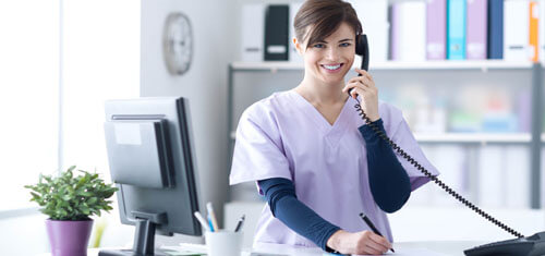 Picture of a receptionist representing Top Plastic Surgeons in beautiful Cancun, Mexico.  The woman has short brown hair, is wearing a hospital smock and is standing at the receptionist desk while smiling at the camera.