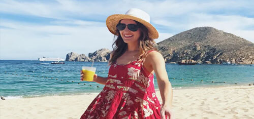 Picture of an attractive woman on a beautiful Cancun beach.  The woman is walking on the beach and is wearing a pink sun dress, straw hat, sunglasses, and is smiling and carrying a drink.